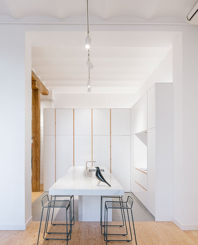 Cocina-con-mobiliario-en-blanco-con-perfilería-de-madera,-isla-de-trabajo-y-office-en-blanco-con-taburetes-en-negro,-escultura-de-pájaro-en-negro,-lumiarias-riel-techo