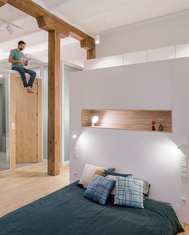 Dormitorio-con-funda-nórdica-azul-marino,-cojines-motivos-geométricos,-módulo-separador-con-estante-escastrado,-armarios-traseros,-vigas-de-madera,-estructura-de-cristal-y-madera,-chico-sobre-puerta