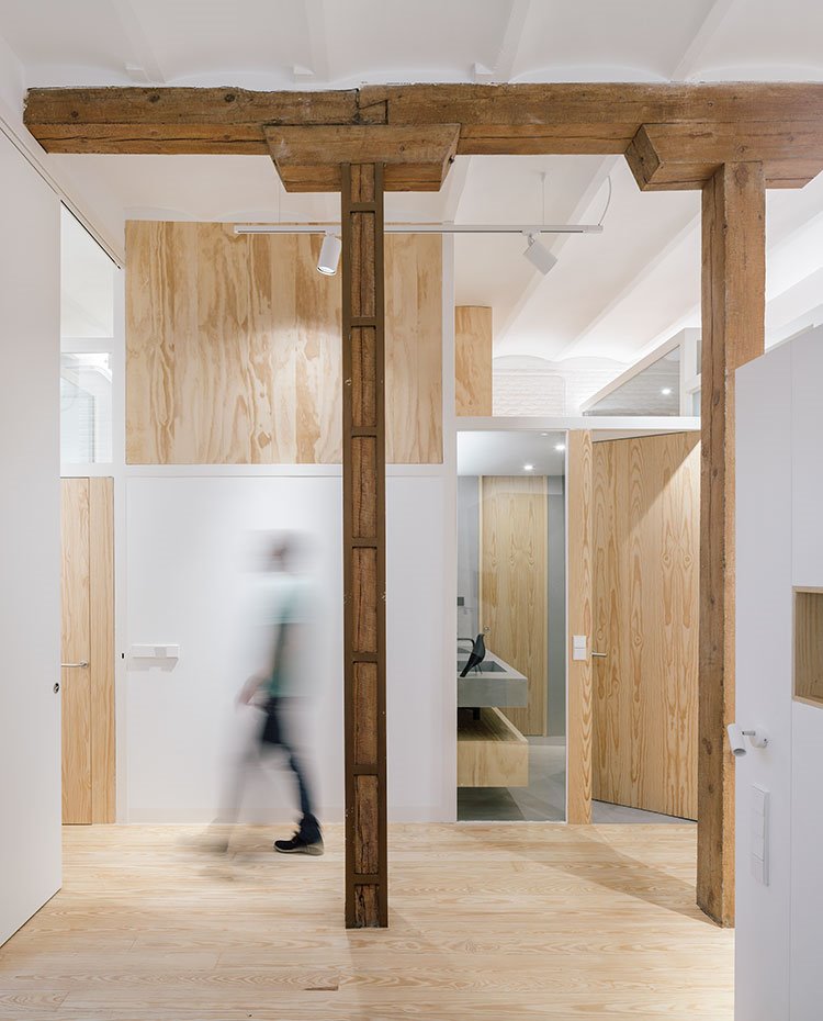 Lateral-dormitorio-con-vigas-y-postes-de-madera,-puerta-corredera-en-blanco,-acceso-a-baño-con-encimera-de-hormigón-y-mueble-bajo-de-madera,-particiones-en-madera-y-cristal