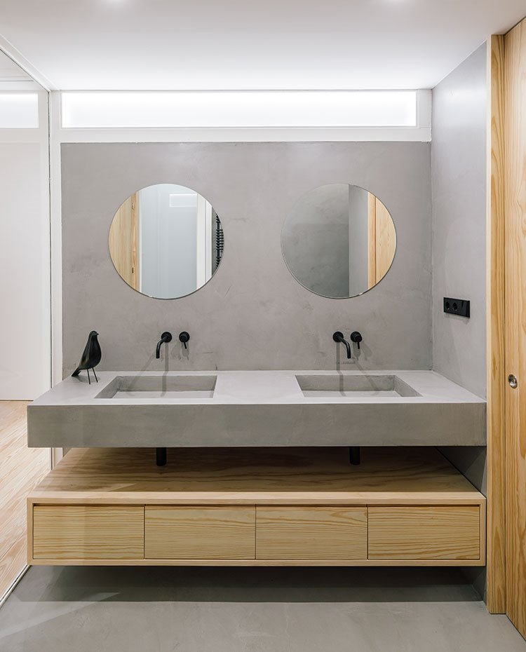 Lavamanos-de-cuarto-de-baño-con-encimera-doble-de-cemento,-grifería-encastrada-negra,-mueble-bajo-suspendido-de-madera-y-suelo-de-hormigón.-Puerta-corredera-de-madera-y-tabique-de-cristal.