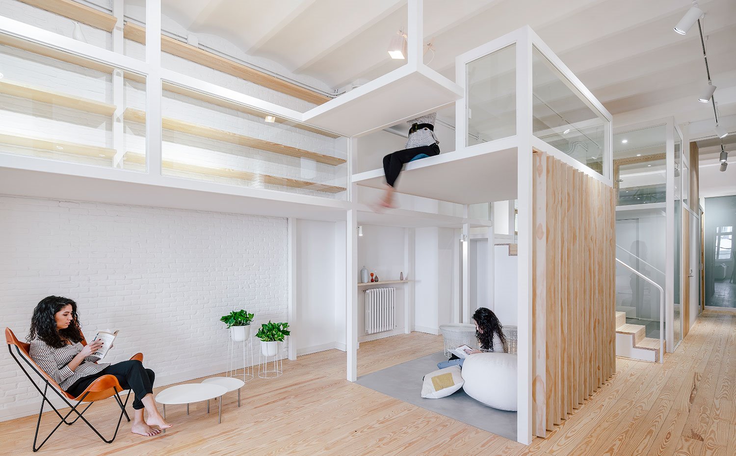 Zona-descanso-con-butaca-marrón-y-estructura-tubular,-mesita-auxiliar-blanca,-revestimiento-con-ladrillo-visto-blanco,-doble-nivel,-baldas-superiores,-estructura-con-alumino-y-madera