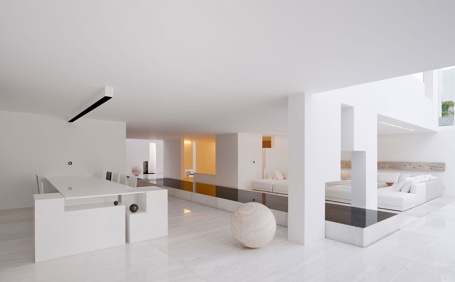 Comedor con estructura rígida en extremo y sillas a juego en blanco, iluminación de techo, esfera de piedra, pasarela decorativa con sobre en negro, salón con sofás en blanco