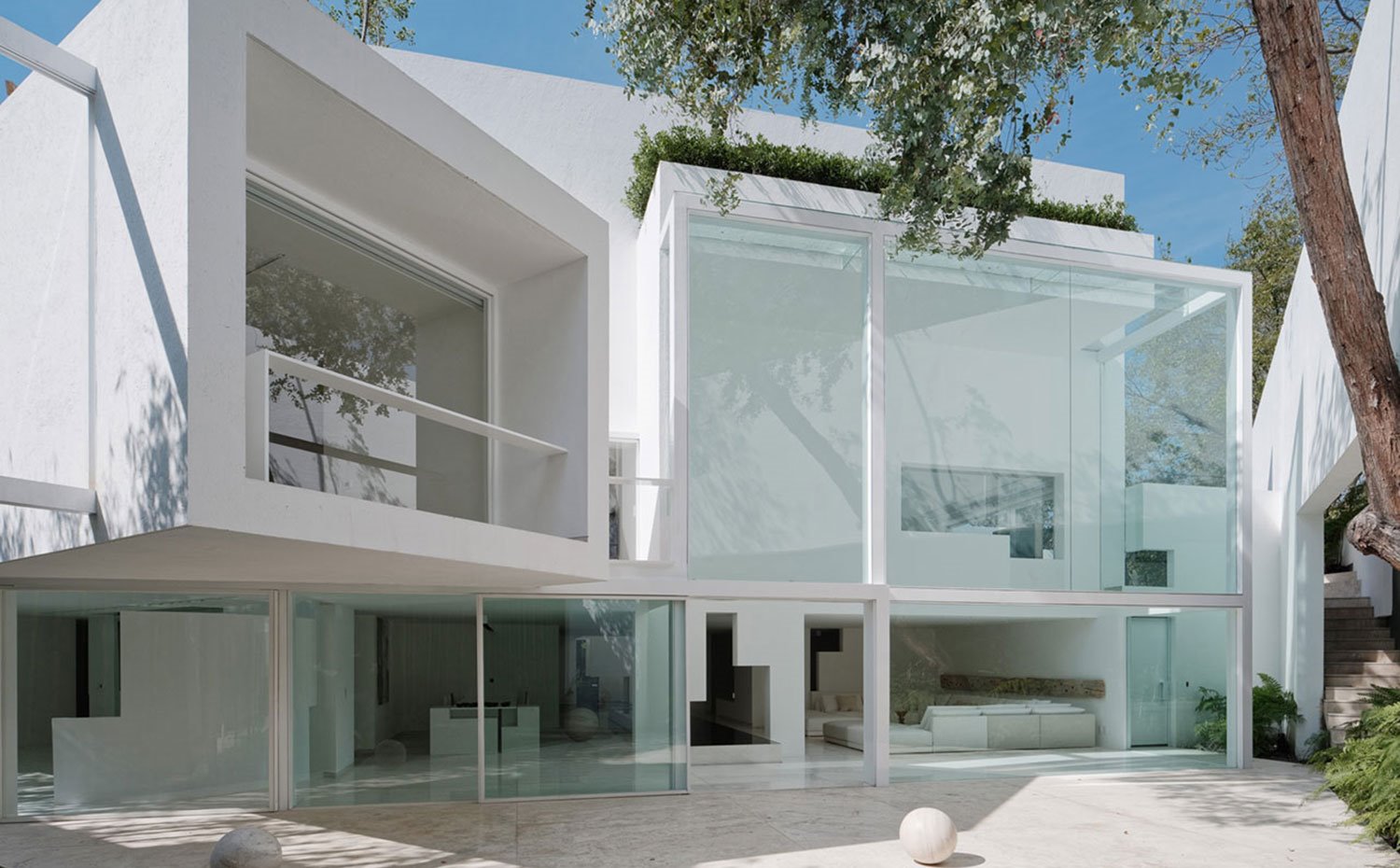 Exterior vivienda con grandes cerramientos de cristal, esfera de piedra, módulos inclinados, vista desde exterior el comedor y el salón interior, vegetación sobre fachada