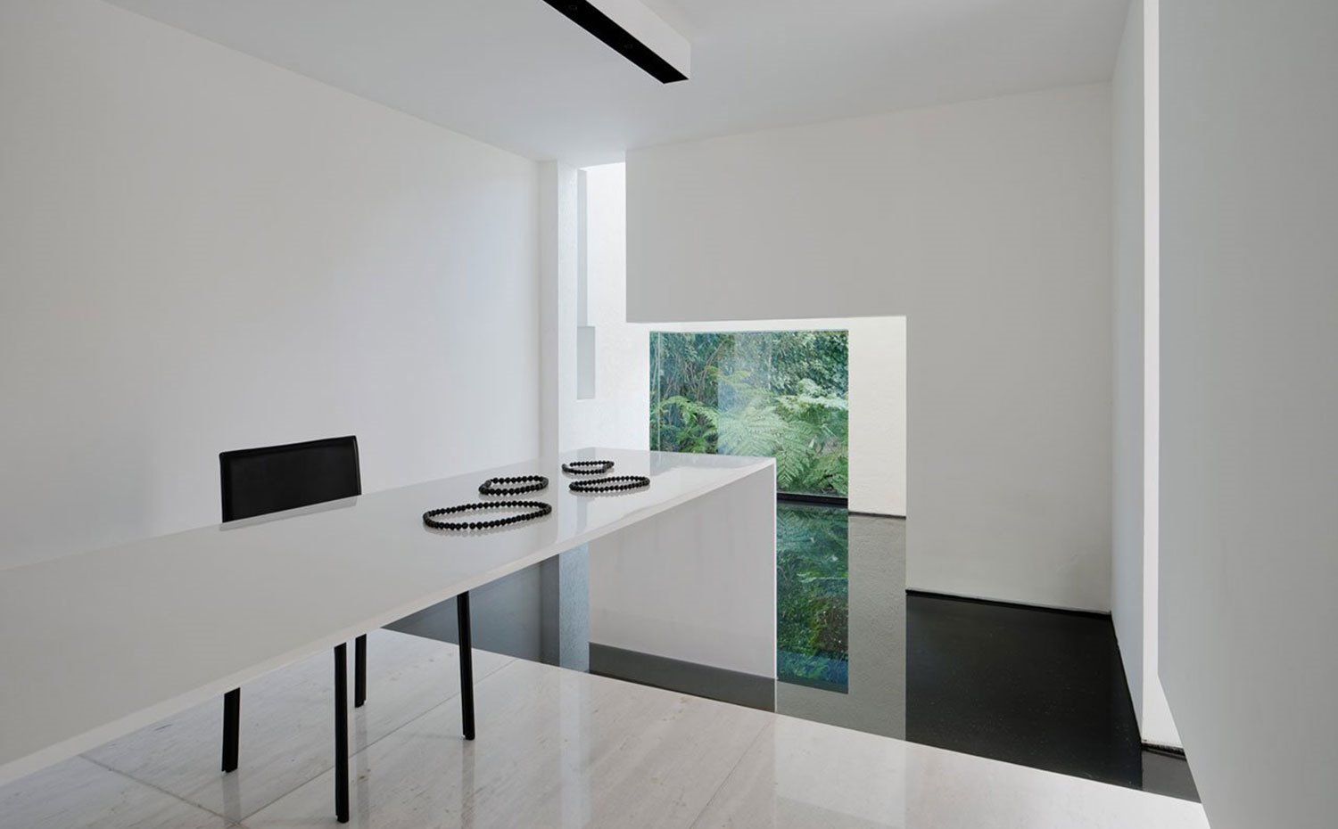 Paredes blancas, suelo de gresite, mesa alargada en blanco, silla negra, base de auga, apertura al exterior con vegetación