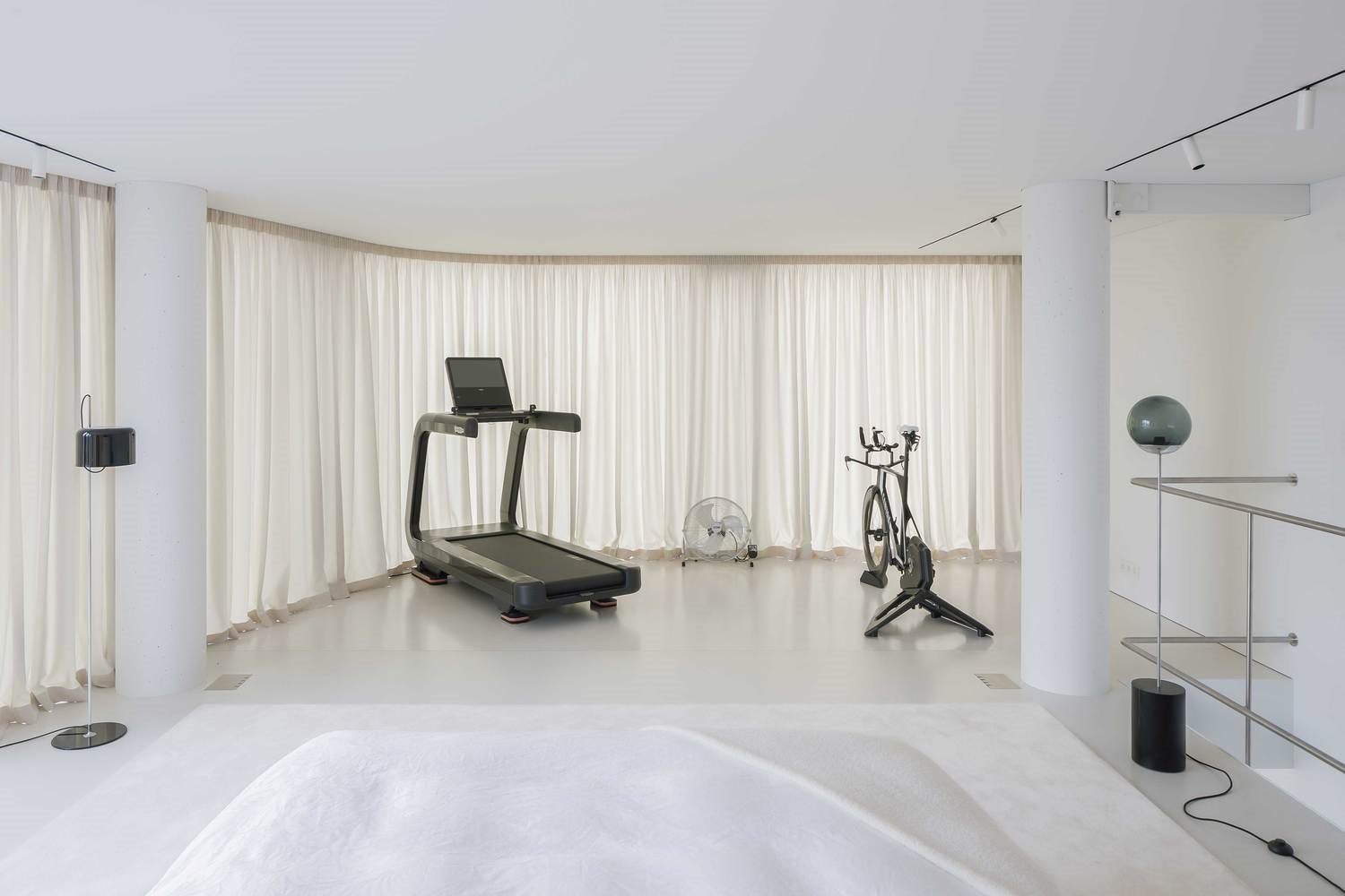 Cama blanca con alfombra en blanco, máquina de correr, bici estática, cortinas corridas