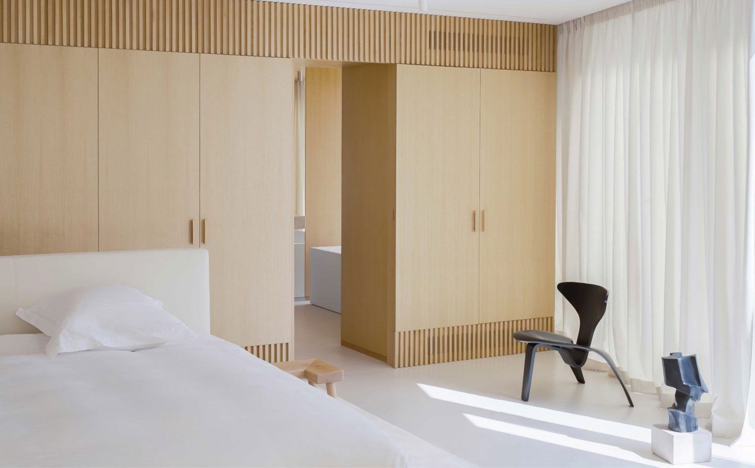 Cama con cabezal blanco, taburete de madera, asiento negro, armarios y revestimiento madera natural, cortinas blancas