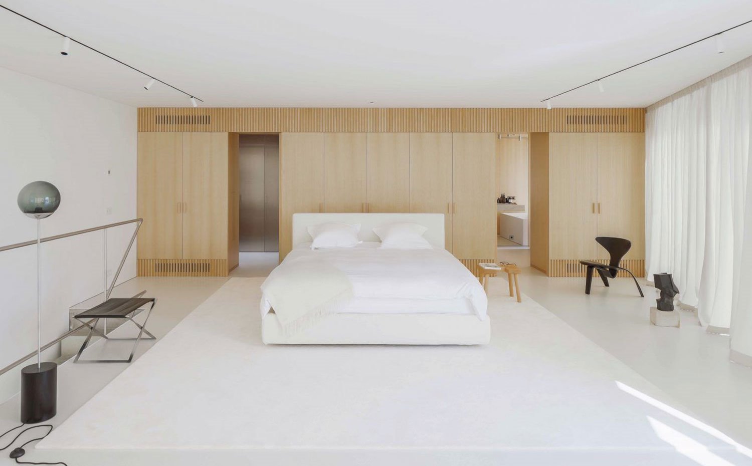 Dormitorio abierto con cama en el centro blanca, armarios de madera natural, taburete de maera, asientos en negro