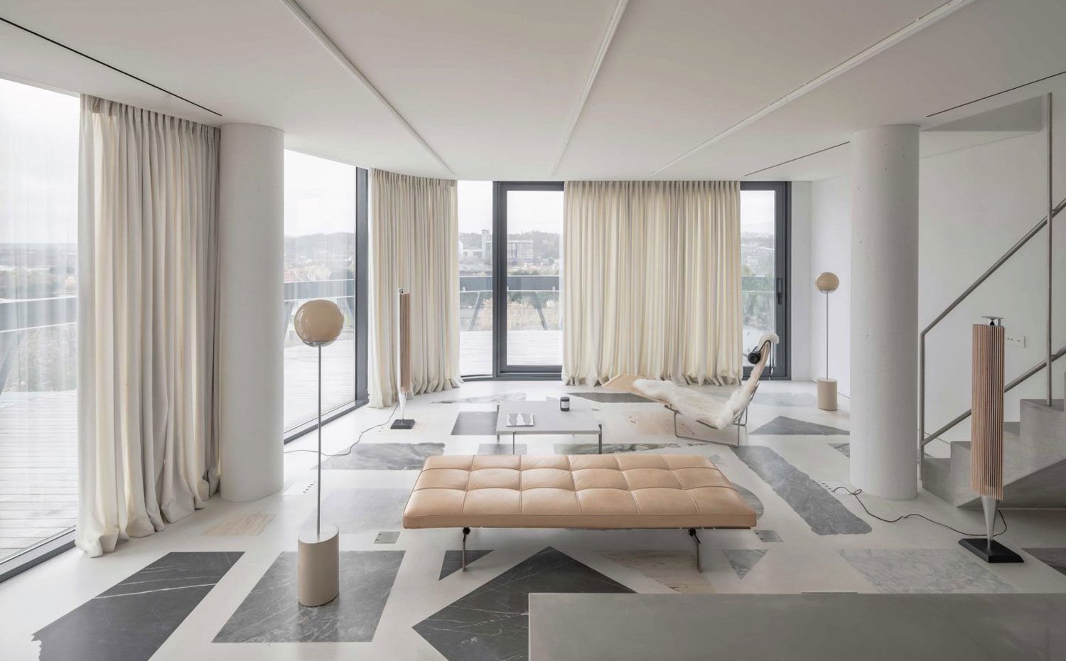 Salón con asiento en capitoné, cortinas frente a cerramientos de crista, lámparas de pie con esfera de cirstal, altavoz de pie, mesita auxiliar y butaca blanca