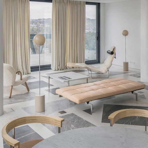 Salón con asiento en capitoné, lámparas de pie con esfera superior, chaise longue beige, butaca con reposabrazos de madera, mesa de mármol, suelo de mármol, cortinas y columnas blancas