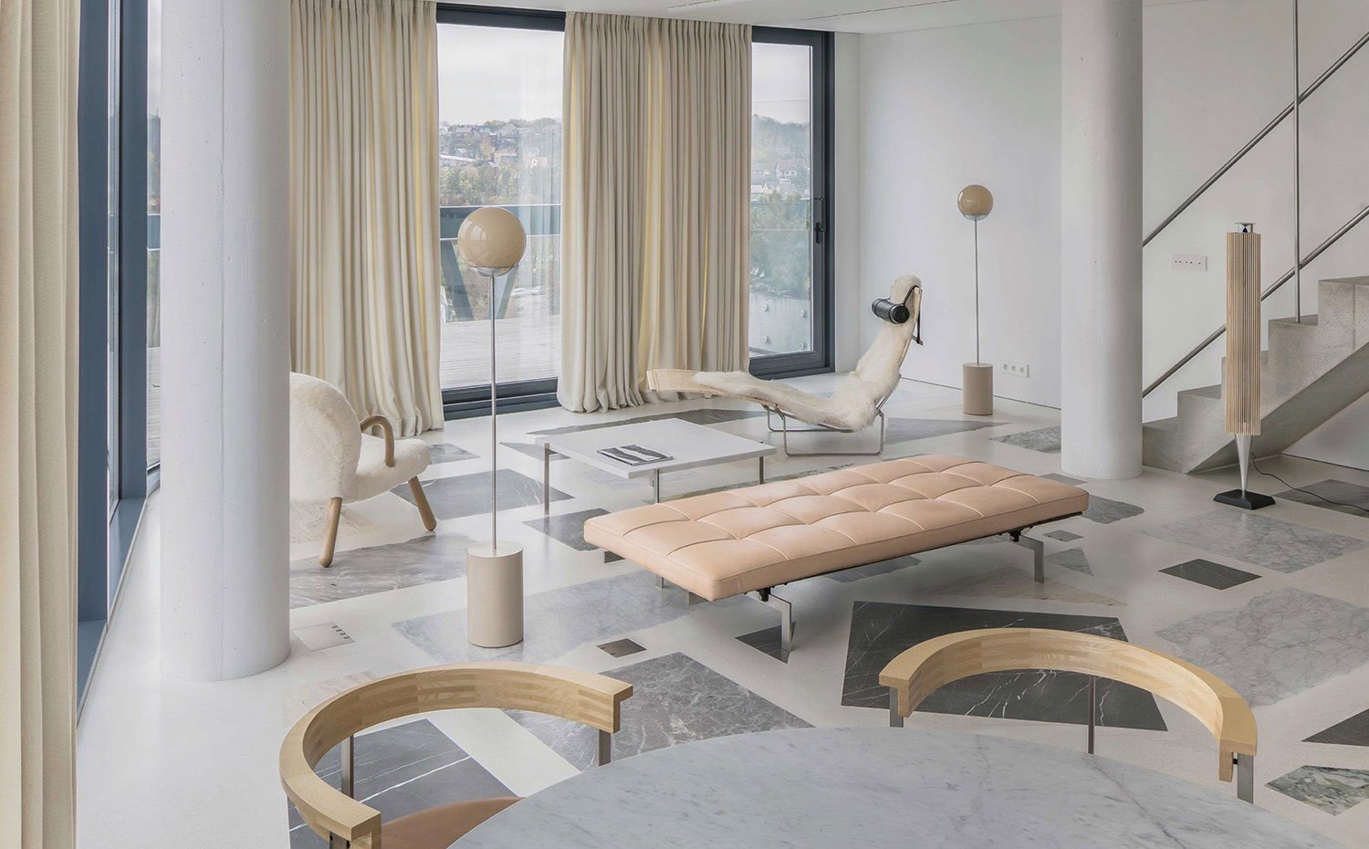 Salón con asiento en capitoné, lámparas de pie con esfera superior, chaise longue beige, butaca con reposabrazos de madera, mesa de mármol, suelo de mármol, cortinas y columnas blancas