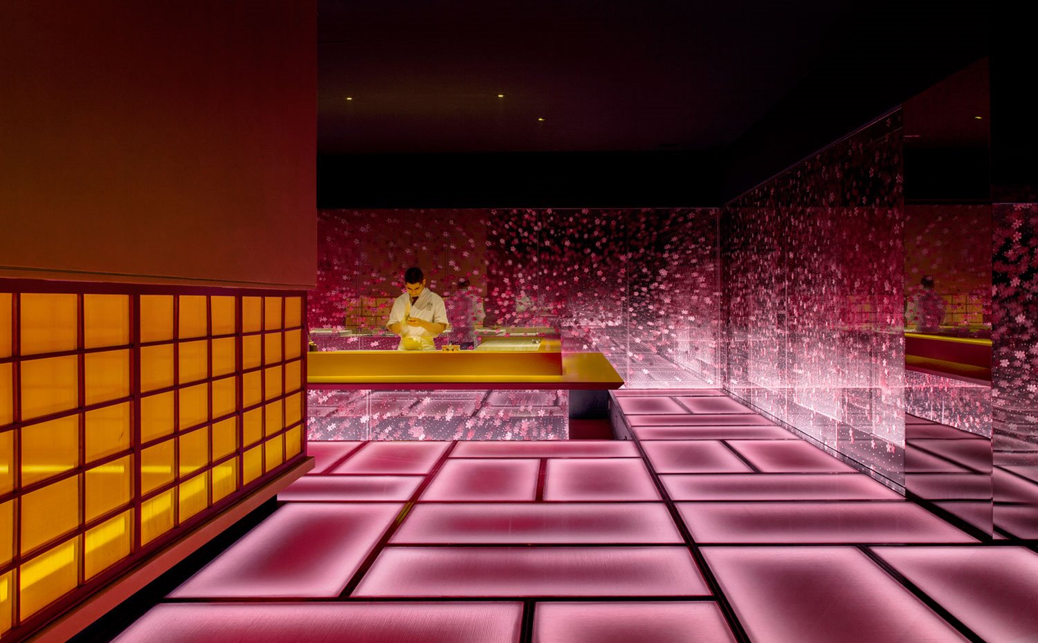 Camarero tras barra iluminada en amarillo y parte inferior de metacrilato rosa. Suelos retroiluminados rosa y metacrilatos con motivos en rosa