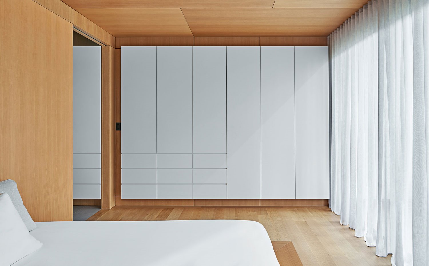 Armarios con frentes lacados en blanco encastrados sobre estructura de madera ntural, cortinas en blanco, cerramiento corredero en madera
