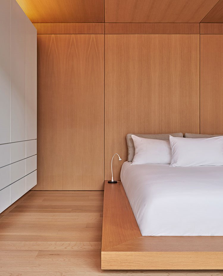 Dormitorio con suelo, paredes y techo de madera, cama sobre estructura baja de madera, armarios almacenaje lacado en blanco, luminarias en lateral madera