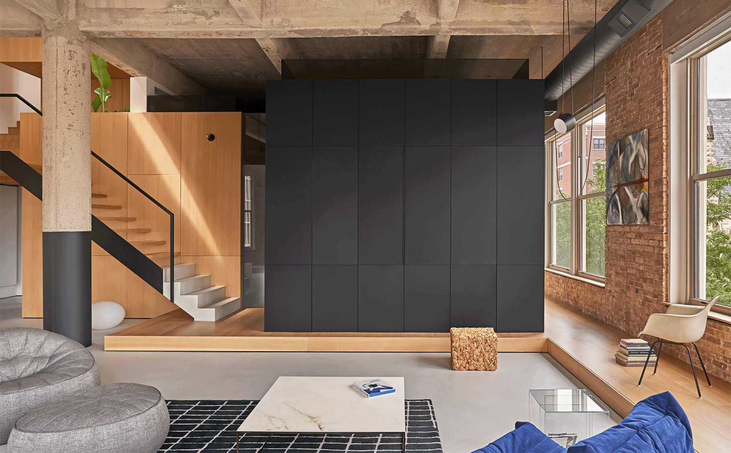 Salón con mueble almacenaje con televisor incorporado en negro, mesa de centro con sobre de mármol blanco, cubierta de hormigón, suelo elevado en madera, silla en blanco, pufs en gris