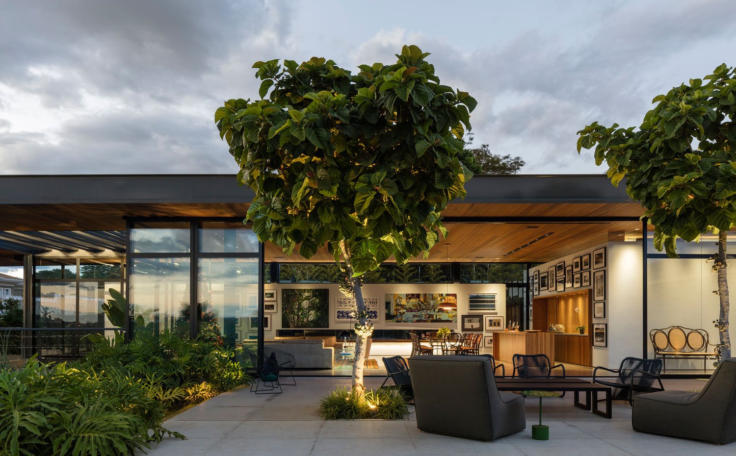 Vista nocturna de exterior vivienda con sillones, butacas y mesa en negro, arbol en centro terraza, cerramiento cristal con sofá en salón, comedor y cocina abierta.