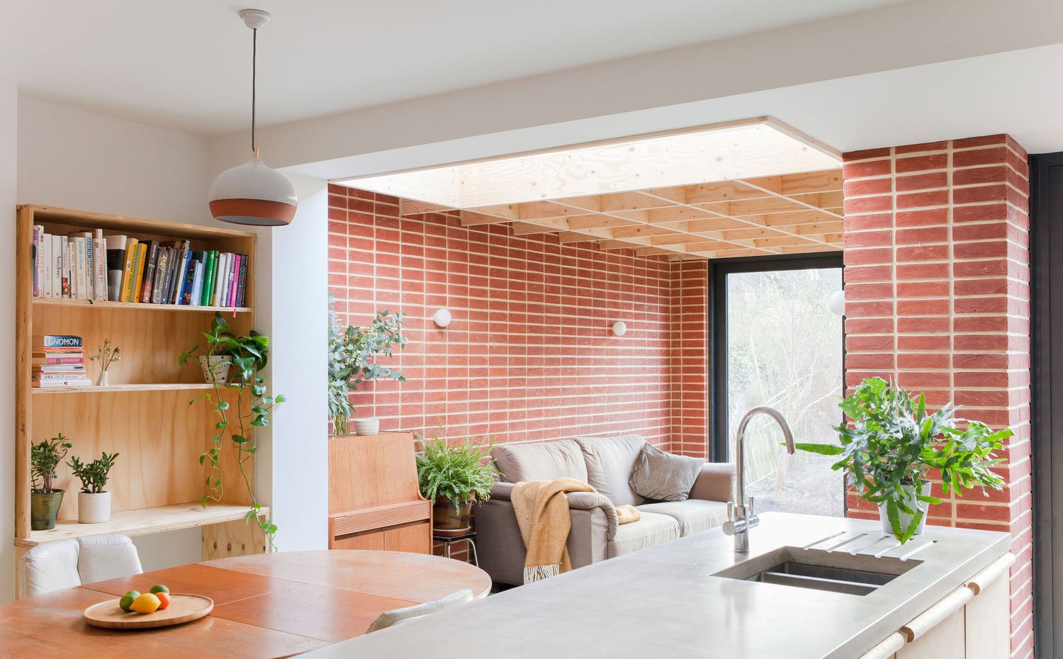 Comedor y salón con estanterías de madera, sofá gris, pared revestimiento ladrillo visto, lámpara suspensión bicolor