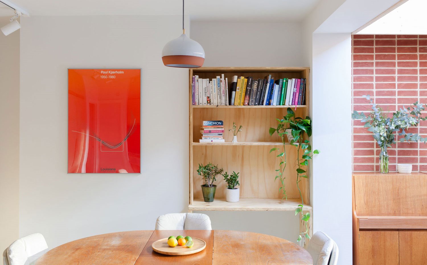 Escritorio y estantería de madera, mesa de comedor circular de madera, fuente de centro en beige, lámina roja en pared, luminaria suspendida en cristal y marrón