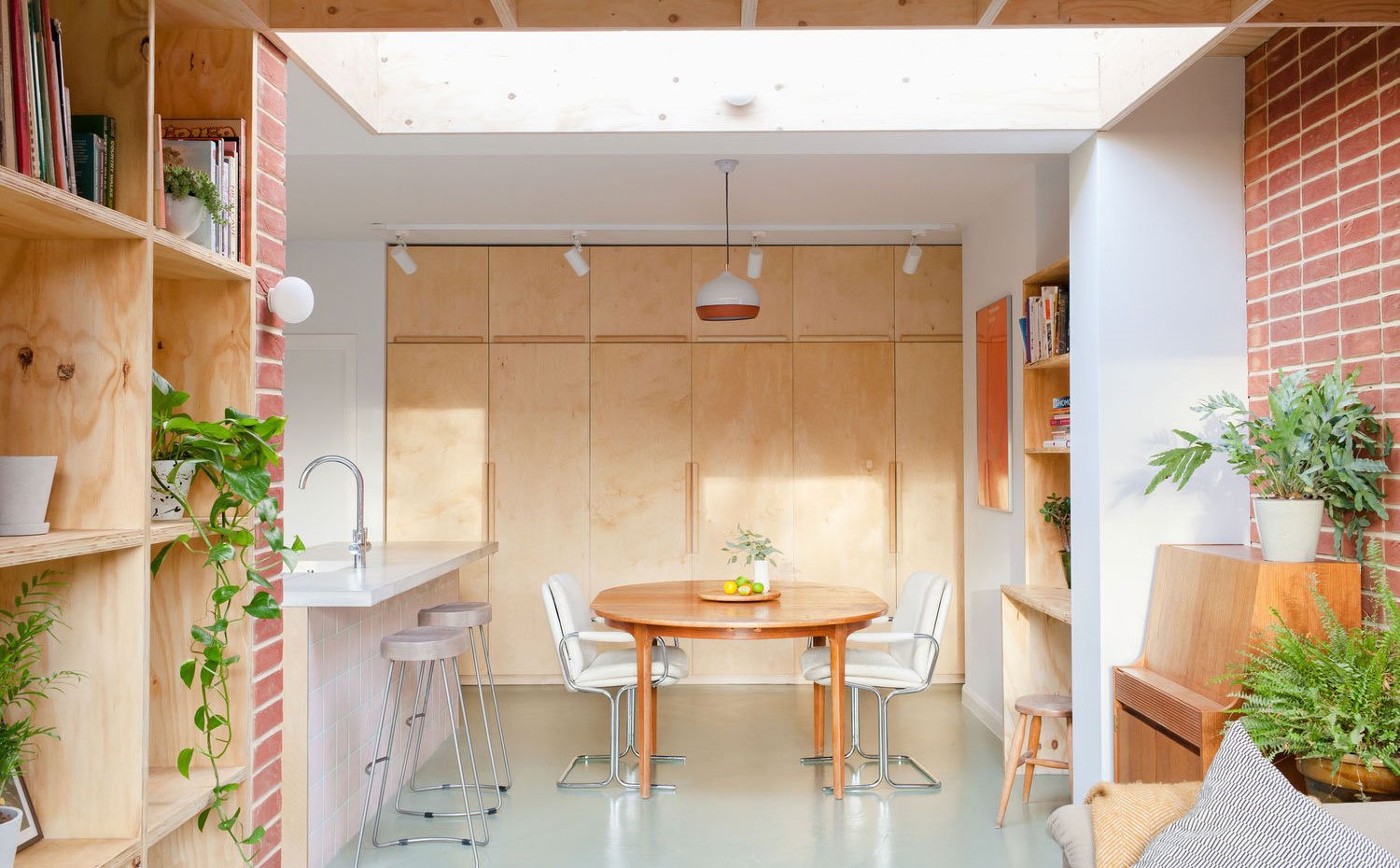 Estantería de madera con plantas, mesa de madera circular, sillas de piel blanca, taburetes grises, armarios de frente madera
