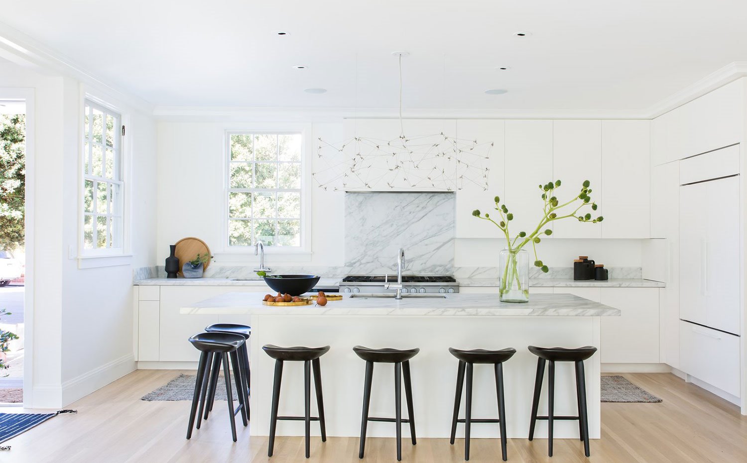 Cocina con salpicadero de mármol, mobiliario blanco, taburete negro, suelo de madera