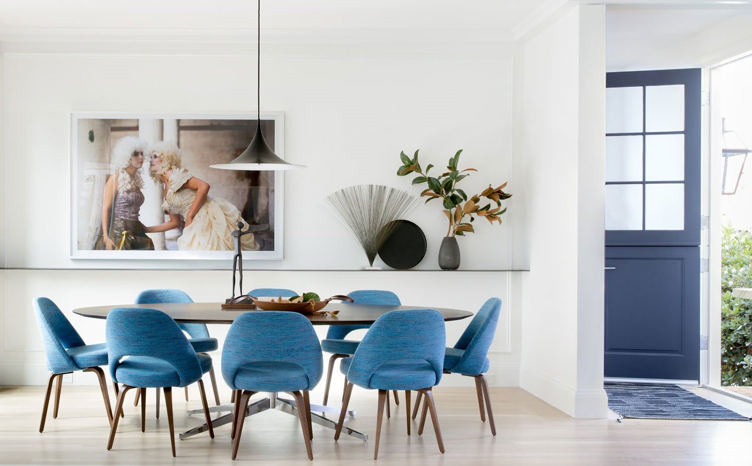 Comedor mesa de madera ovalada, sillas tapizadas en azul, escultura centro de mesa, balda con jarrones y elementos decorativos, cuadro de inspiración victoriana, puerta azul