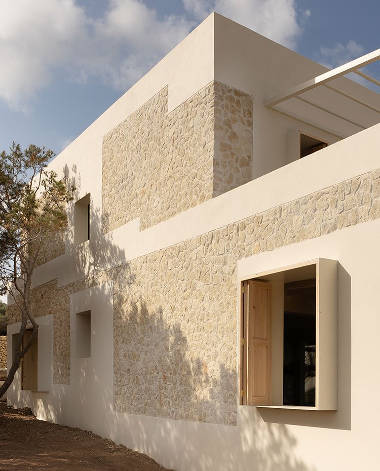 Vista lateral vivienda con árboles que le dan sombra, fachada de piedra y hormigón y ventanas cuadradas con porticones de madera
