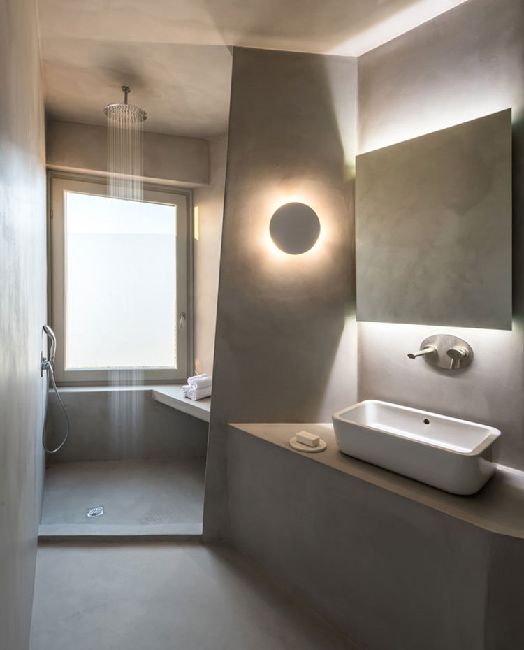 Baño totalmente de hormigón con lavamanos de cerármica y grifería de acero, iluminación indirecta en espejo y aplique y rociador en el techo de la ducha