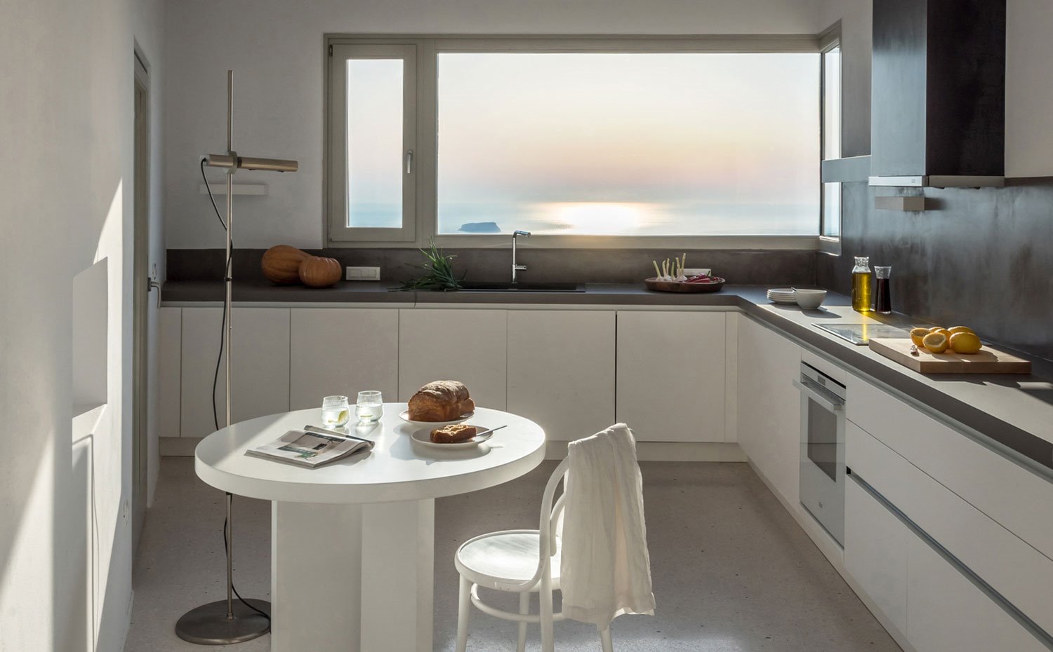 Cocina con mobiliario inferior en blanco con tiradores integrados, salpicadero y encimera en gris, electrodomésticos en acero, mesa y silla de office en blanco y lámpara de pie de líneas depuradas