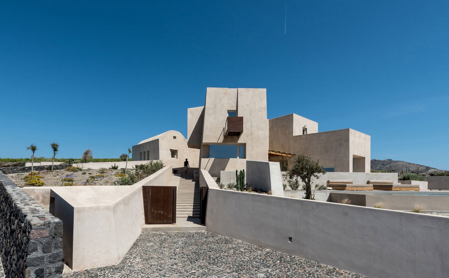 Entrada con escaleras, edificio de hormigón, murete de piedra, suelo de cantos rodados