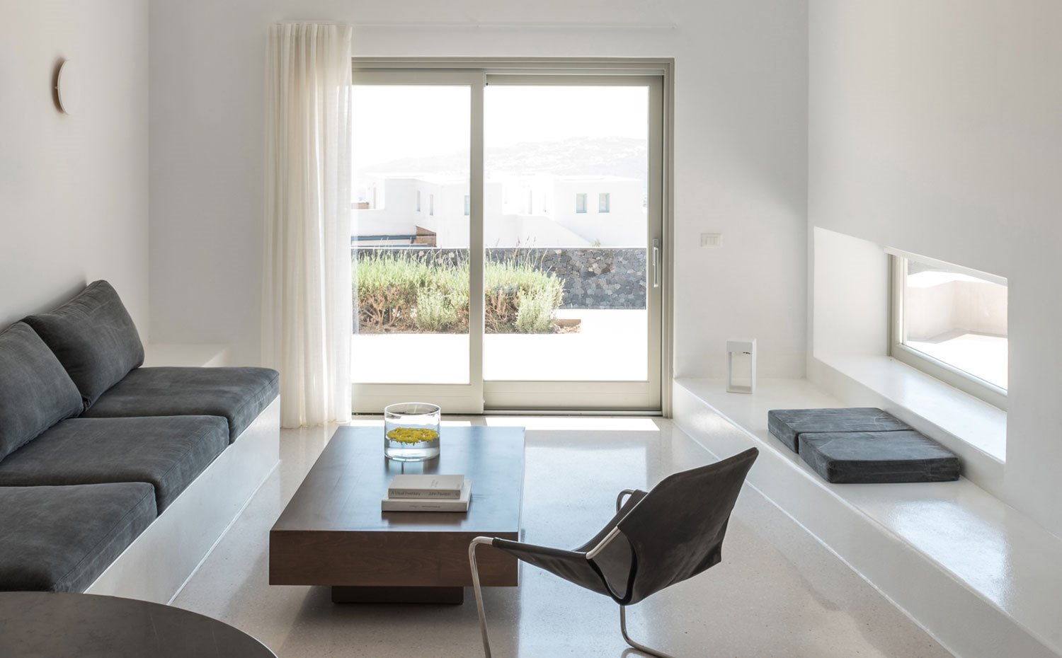 Salón con mesa de centro de madera, sofá con estructura de cemento y cojines en negro, butaca de piel y acero y cortinas blancas 