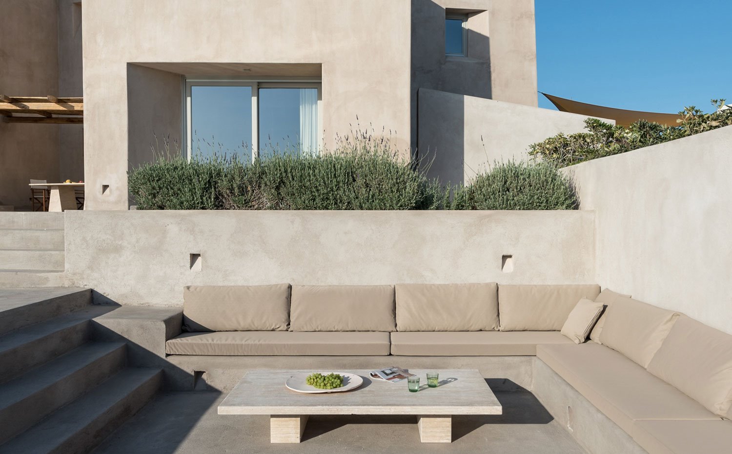 Salón exterior en hormigón y yeso, con banco corrido esquiner con cojines del mismo tono, vegetación silvestre, mesa de centro baja de madera