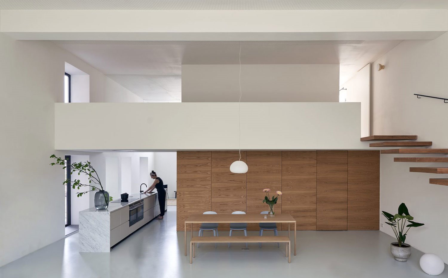 Cocina y comedor en espacio abierto a doble altura con escaleras en voladizo, lámpara suspendida a gran altura, jarrón decorativo sobre encimera mármol de isla alargada de cocina