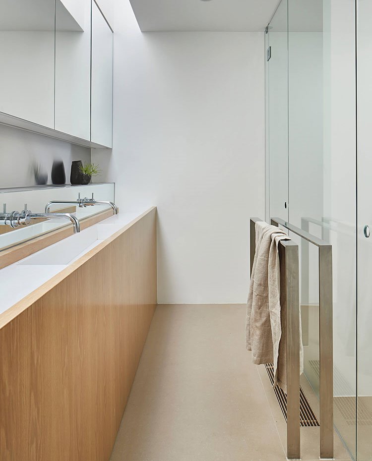 Cuarto de baño con doble zona de lavamanos, armario superior con espejo y parte inferior en madera natural, toallero de pie de acero, cerramiento tabique de cristal 