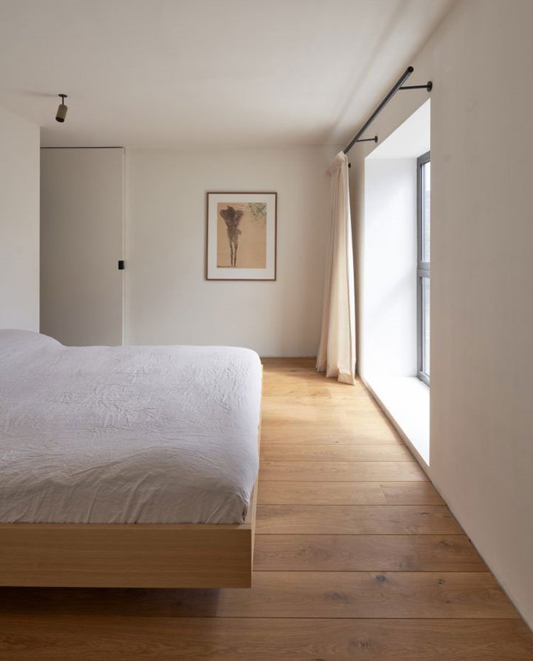 Dormitorio con estructura suspendida en madera, lencería de cama blanca, suelo de madera, cuadro de tonos tierra en pared, cortinas de tonos ocre, foco de techo, puerta lacada blanca