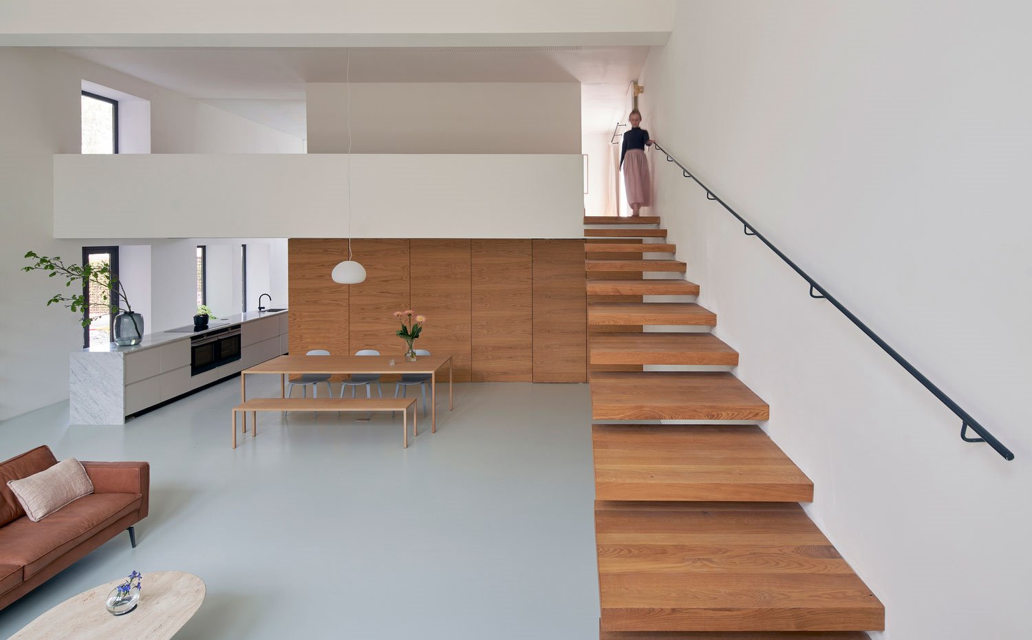 Escaleras en voladizo de madera, pasamanos negro, armarios de almacenaje de madera, sofá marrón, cocina en isla