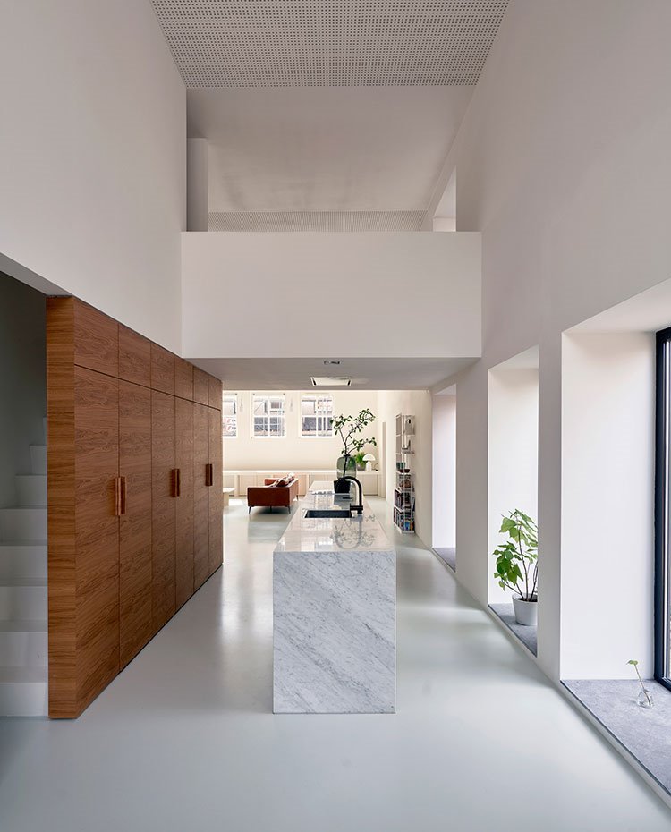 Espacio de doble altura con escaleras blancas hacia el piso superior, frentes de armarios en madera, encimera y lateral isla de mármol, balcón piso superior
