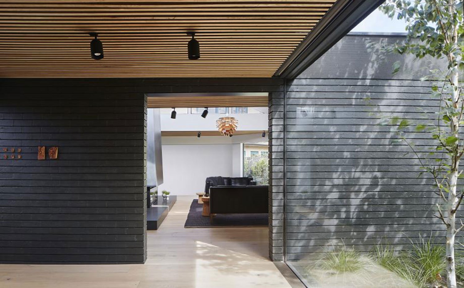 Interior vivienda con cerramiento acristalado y vistas a vegetación exterior, techo con listones de madera y focos en negro, salón con chimenea y sofás negros