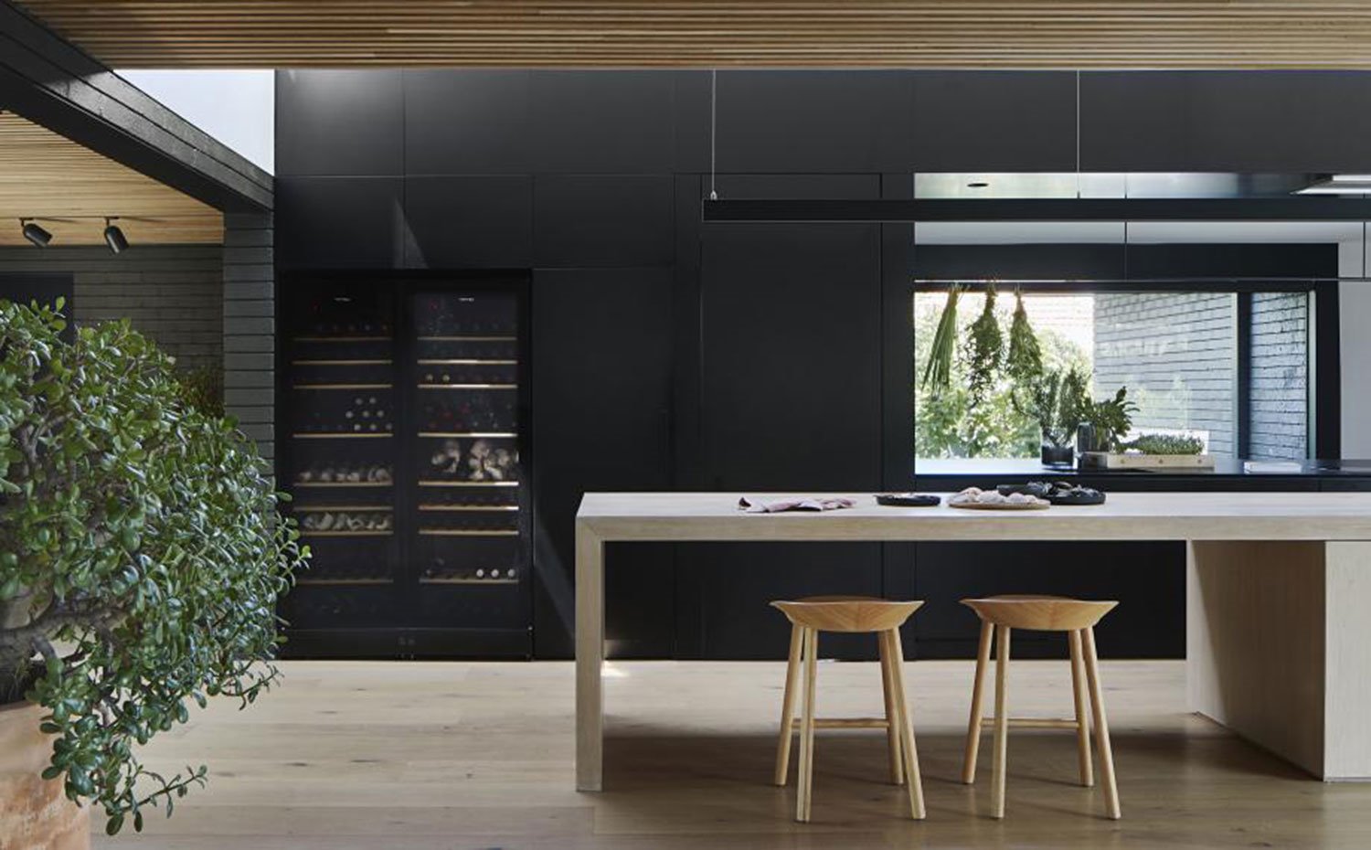 Isla de cocina y taburetes en madera natural, luminaria suspendida alargada, techo de listones de madera, frente de almacenaje lacado en negro