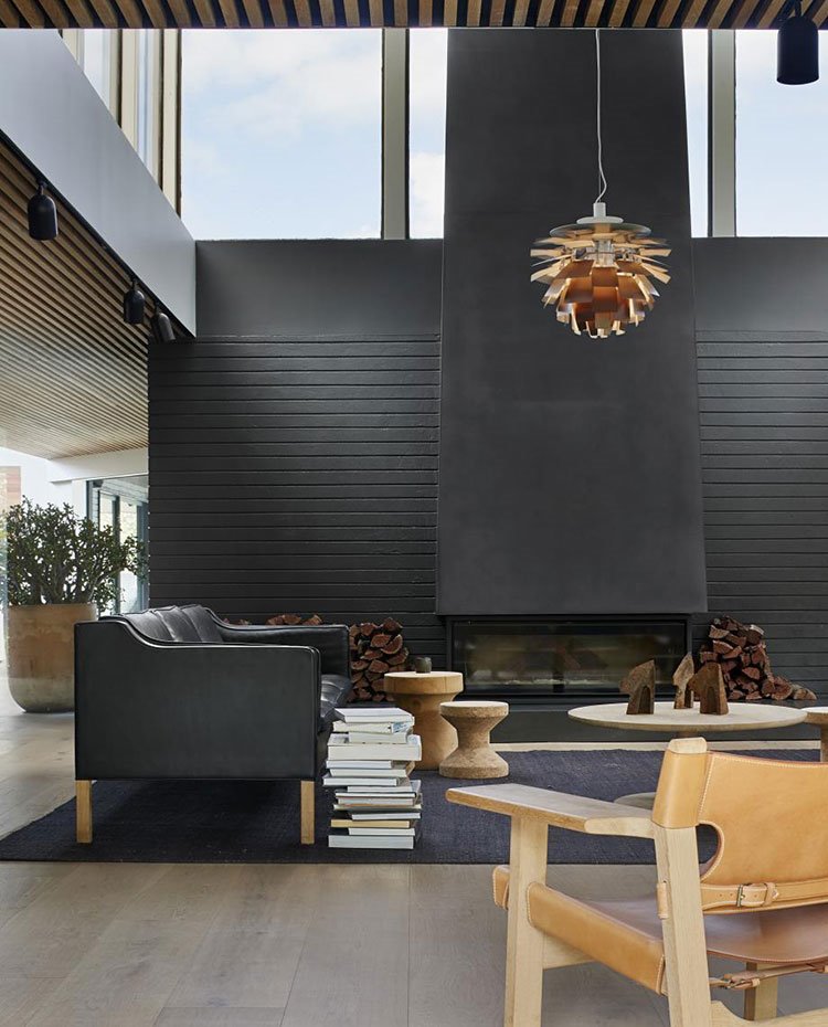 Salón a doble altura, sofá negro, mesas y taburetes en madera, silla en madera y piel, alfombra gris antracita, suelo de madera, chimenea e hierro