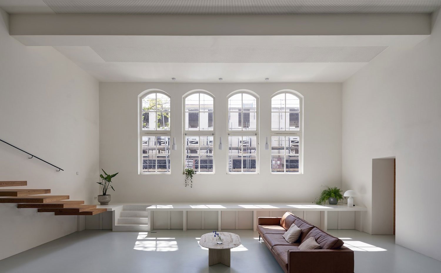Salón con sofá marrón, cojines beige, mesa de centro con detalle cristal, ventanas a gran altura, escaleras en voladizo en pared de madera, estructura elevada bajo ventanas