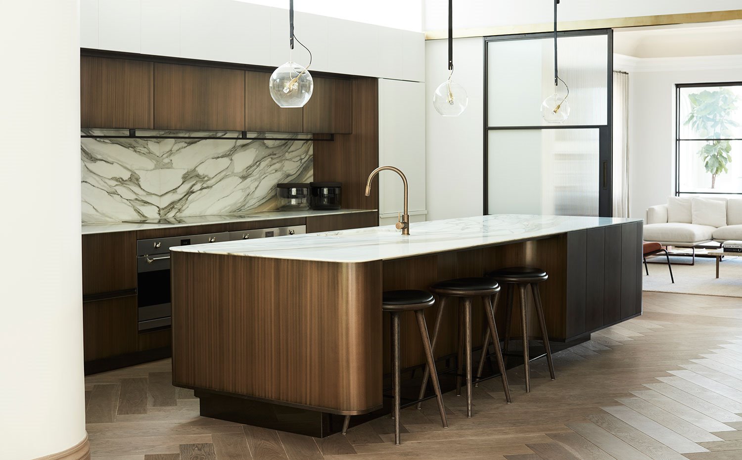 Cocina con frentes de armario de madera oscura, salpicadero y encimera de mármol, isla con office con tres taburetes en acabado negro y bronce, pavimento de madera
