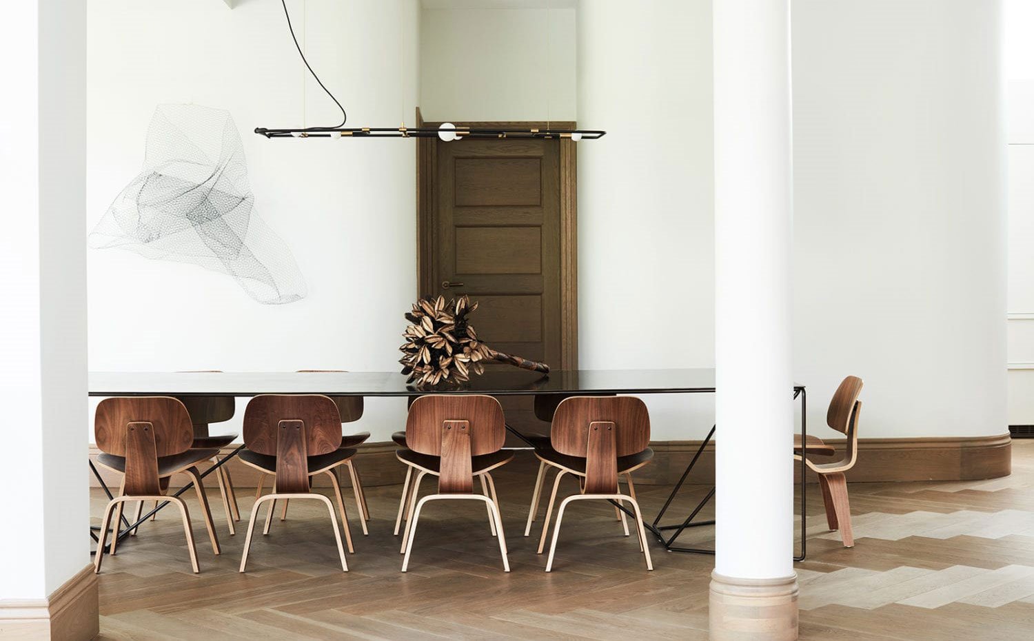 Comedor con columnas y pilares, mesa con encimera de cristal y base de acero negro, sillas de madera, luminaria suspendida de líneas puras, suelo de madera en espiga, centro de mesa de bronce