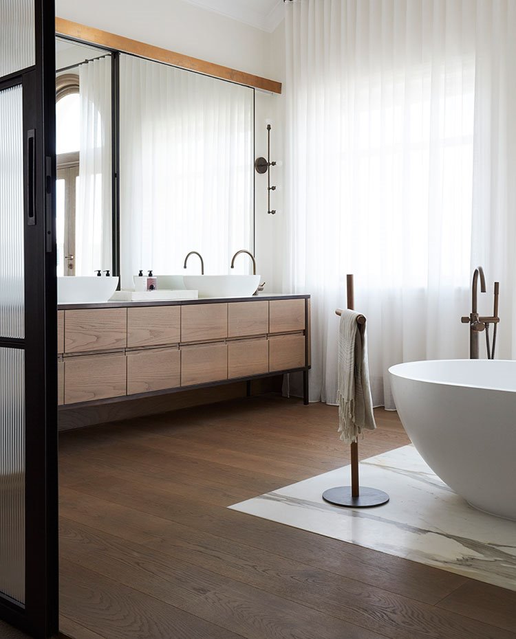 Cuarto de baño con bañera exenta, suelo de madera y mármol, mueble de lavamanos suspendido de madera, doble lavamanos, toallero de pie, puerta corredera, cortinas blancas 