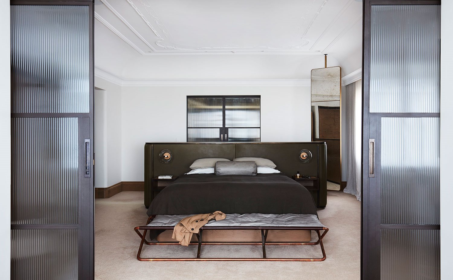 Dormitorio principal con cabecero envolvente tapizado de piel y bancada a pie de cama en gris y cobre, gran espejo vertical apoyado en techo, puertas correderas con cristal 