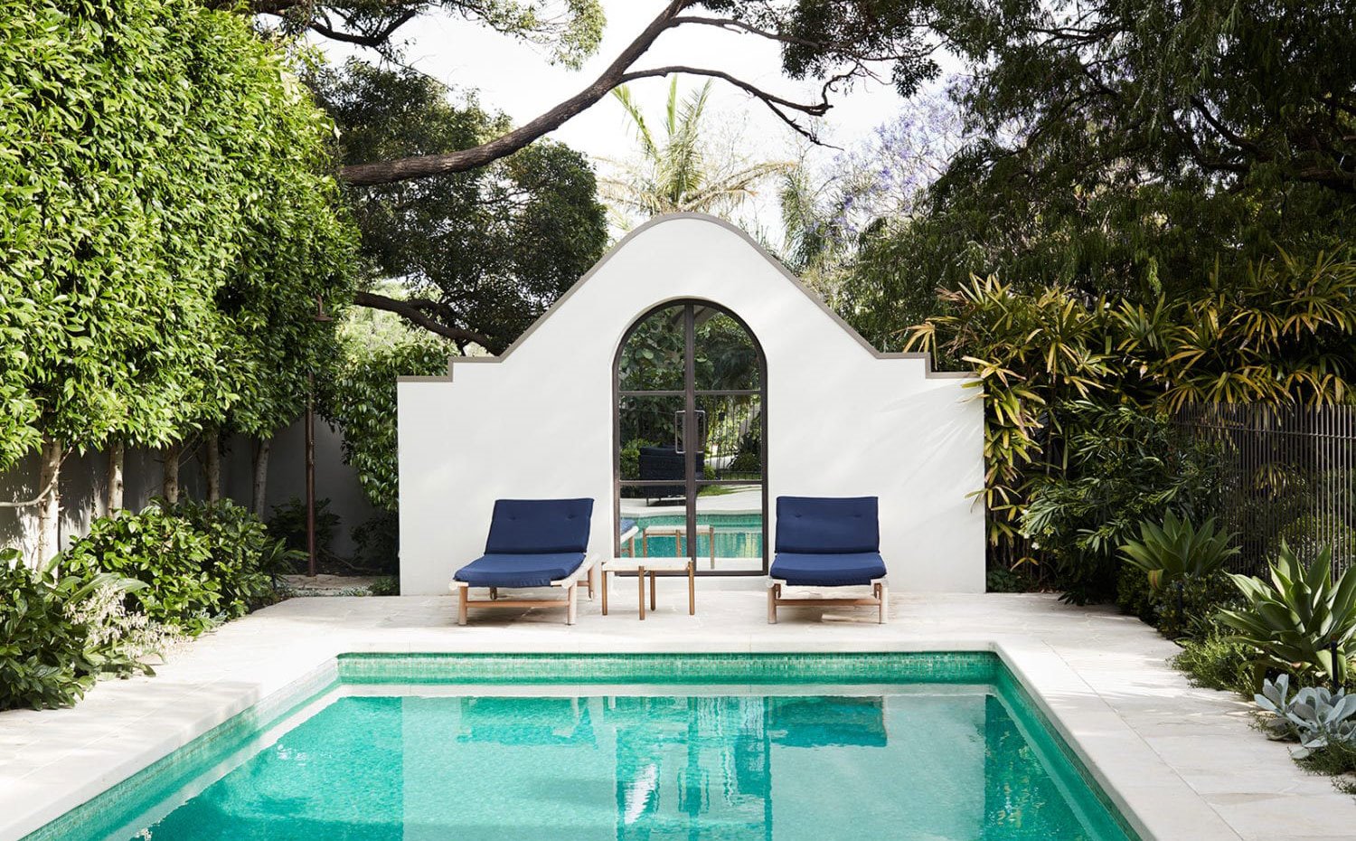 Piscina exterior rodeada de vegetación, hamacas con estructura de madera y cojín azúl, caseta de pisicna en blanco y puerta de efecto espejo