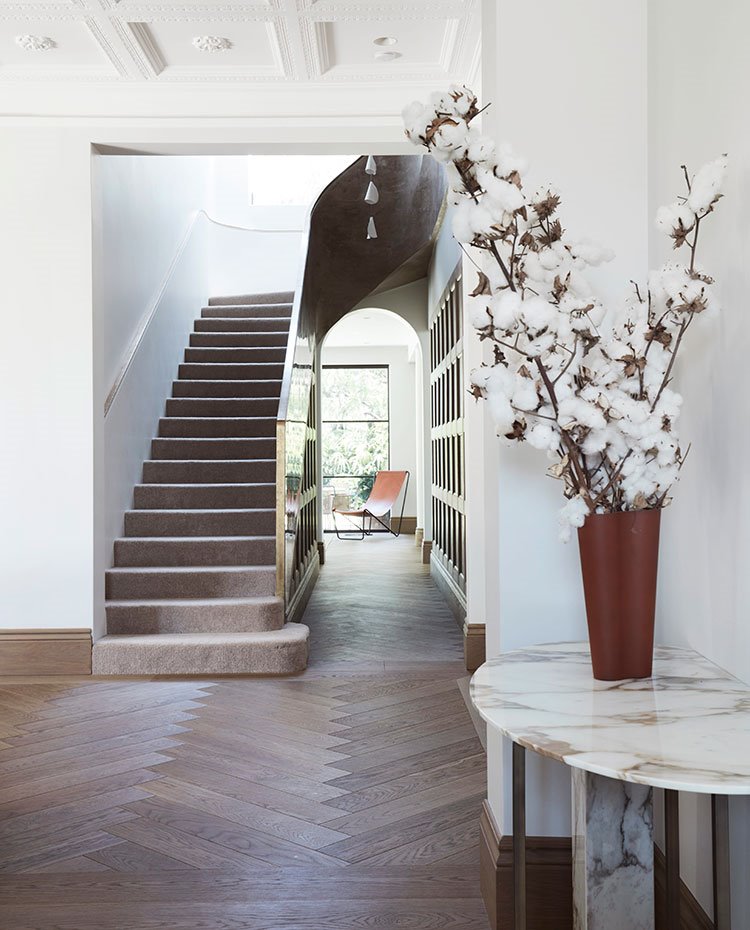 Zona de paso con pavimento de madera en espiga, mesa decorativa en mármol con jarrón y flores de algodón, revestimiento pared en madera con molduras y escaleras con moqueta
