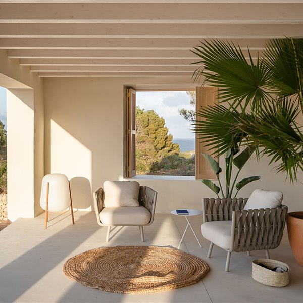 Porche posterior de la vivienda con alfombra de rafia redonda, un gran macetero con planta tropical, butacas en tonos crudos y moderna luminaria de suelo