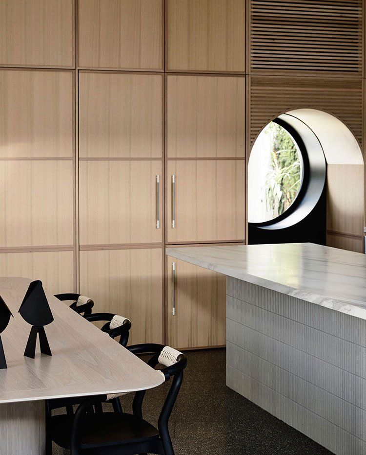 Zona de almacenaje cocina con frente de armarios de madera, mesa comedor con sillas en negro, isla de trabajo con encimera en voladizo, ventana circular. Zona de almacenaje con frente de armarios de madera, mesa comedor con sillas en negro, isla de trabajo con encimera en voladizo, ventana circular