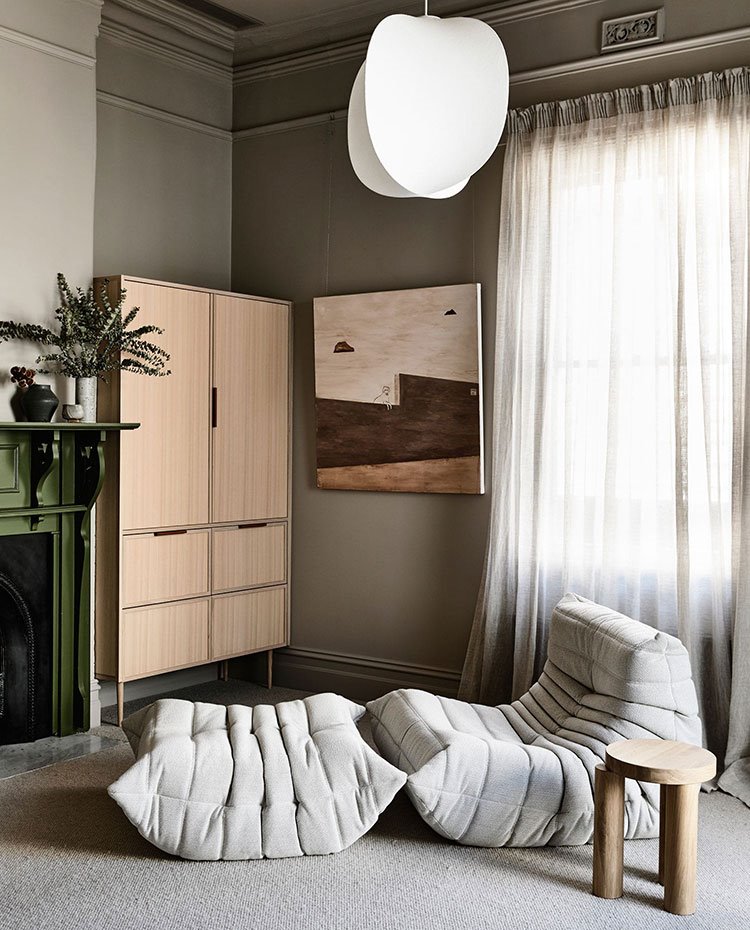 Zona de lectura con asiento y puff informales, taburete madera, mueble de maera junto a chimenea cuadro en pared y cortinas frente a ventana