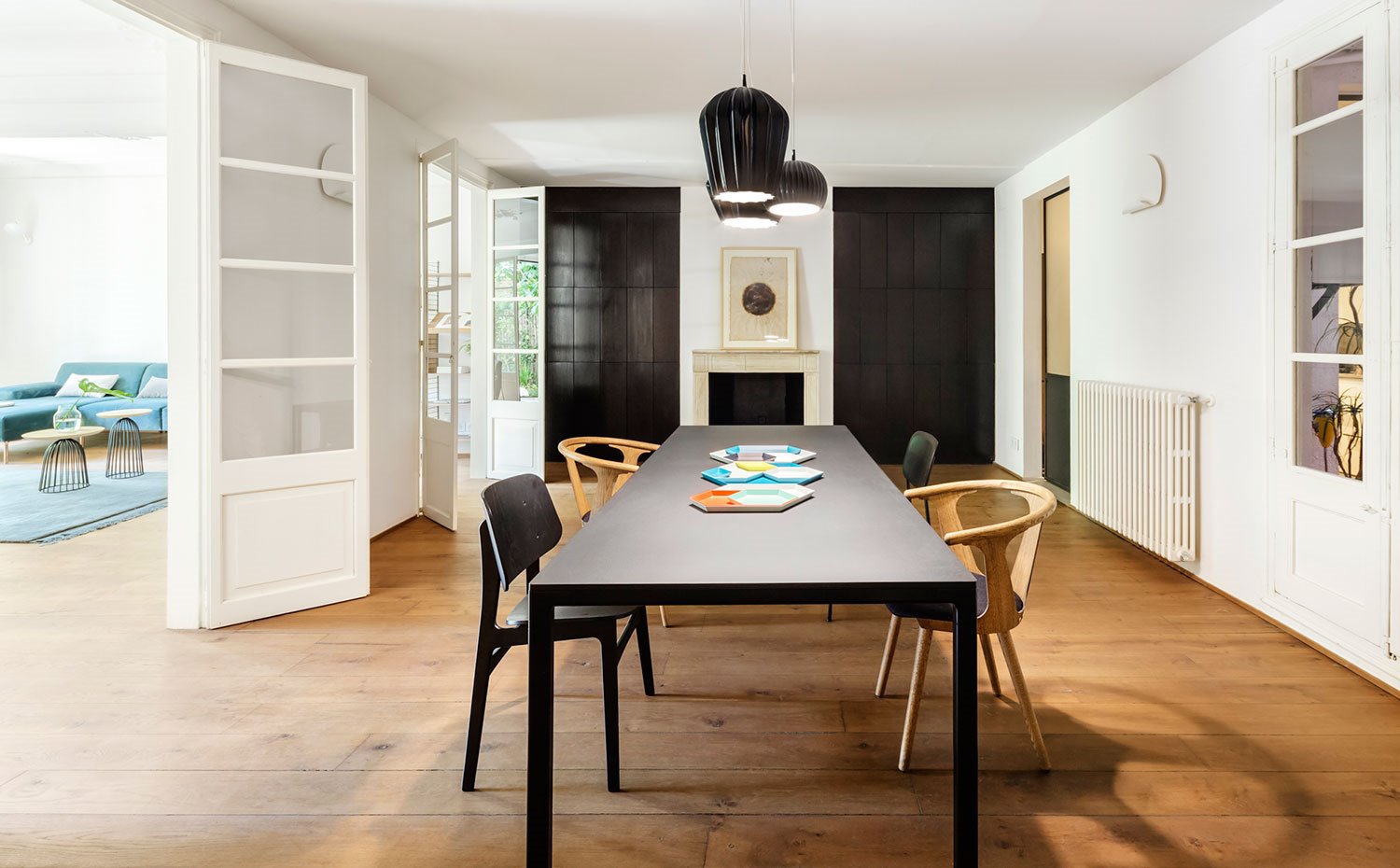 Comedor, mesa de centro negra, sillas de madera, luminarias suspendidas negras, pavimento de madera, puertas de madera y cristal de acceso a estancia, radiador pared blanco