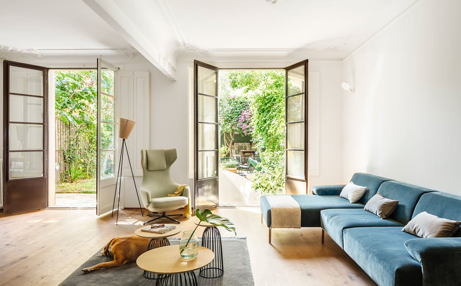 Salón con grandes aperturas a jardín exterior, sofá azul con cojines grises, mesillas auxiliares de acero y madera, alfombra negra, butaca verde, luminaria de suelo con pantalla cónica